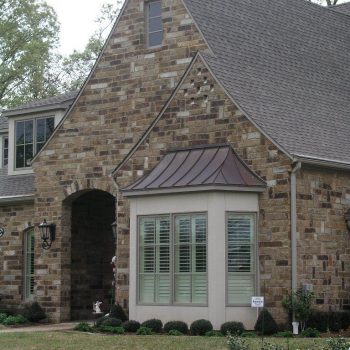 Brick house with steep roof, arched entryway, bay window, and small flowerbed in the front yard.