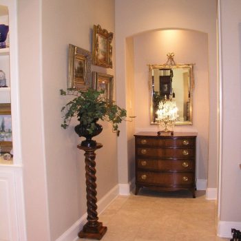 Narrow hallway with framed art, a potted plant on a pedestal, and a dresser with a mirror and lamp at the end.
