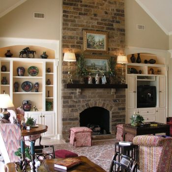 A living room with a tall stone fireplace, built-in shelves, and traditional furniture arranged around a patterned rug.