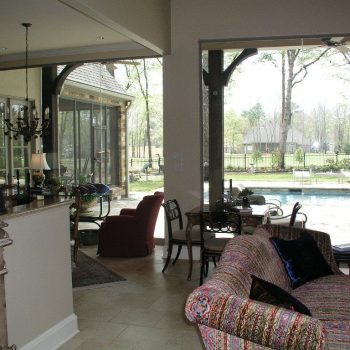 Living room with patterned furniture, glass doors open to patio and pool, garden and trees visible outside.