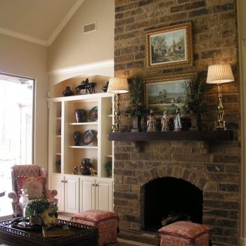 Living room with a tall stone fireplace, framed art, lamps, shelves with decor, and two ottomans on a patterned rug.