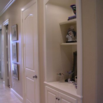 Hallway with beige walls, a closed door, framed pictures, and a built-in illuminated white display cabinet with shelves.