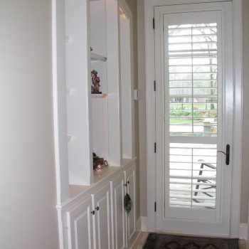 White built-in shelving unit with decor items next to a glass door with white blinds leading to an outdoor area.