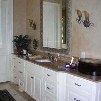 Bathroom vanity with white cabinets, large framed mirror, wall sconces, and various toiletries on the countertop.