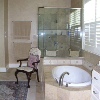 Bathroom with a glass-enclosed shower, built-in bathtub, a cushioned chair, tiled floor, and towels on a rack.