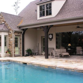 Covered patio with outdoor seating and ceiling fan overlooks a swimming pool beside a stone and stucco house.
