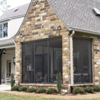 A house with a screened-in porch featuring stone exterior walls and a sloped roof, surrounded by a landscaped yard.