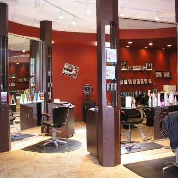 Modern hair salon interior with styling stations, chairs, mirrors, hair products, and photos on red walls.