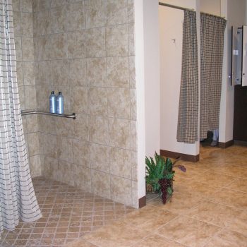 Tiled shower area with two bottles, a grab bar, two curtained changing stalls, and a decorative plant on the floor.