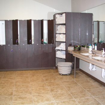 Locker room with open lockers, shelves of rolled towels, sinks with soap dispensers, and a basket for used towels.