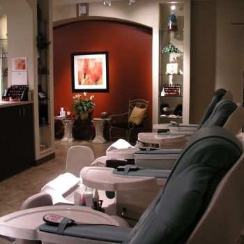 Rows of empty pedicure chairs face a cozy, softly lit spa interior with a red accent wall and framed artwork.