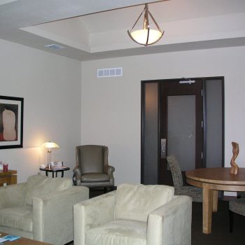 A waiting room with beige chairs, a round table, artwork on the walls, and a closed door in the background.