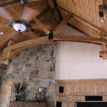 Exposed wooden beams and stone wall in a living room with a vaulted ceiling and ceiling fan.