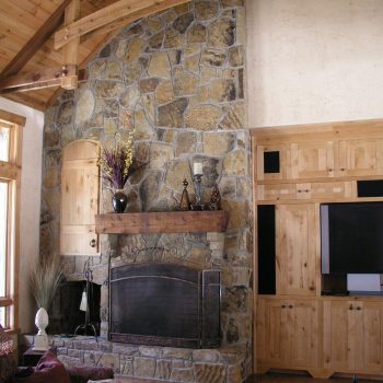 Living room with a tall stone fireplace, wood mantel, built-in wooden cabinets, and exposed wood ceiling beams.