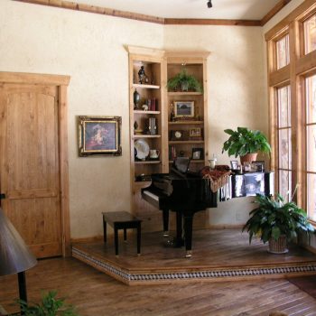 A grand piano sits on a raised platform by tall windows, surrounded by plants and wooden bookshelves in a sunlit room.