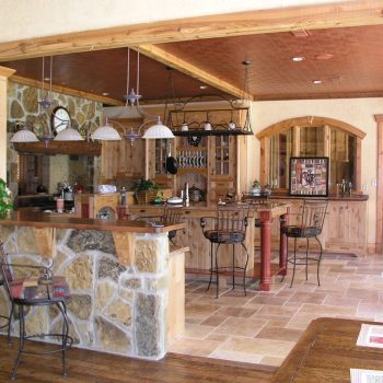 Rustic kitchen with stone and wood accents, bar seating, pendant lights, and tiled floor.