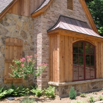 House exterior with stone, brick, and wood siding, large window, and landscaped garden with small flowering tree.