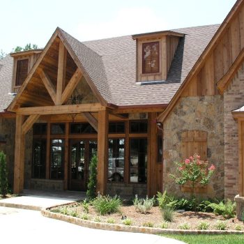 A rustic house with stone and wood exterior, large windows, peaked roof, and landscaped front yard.