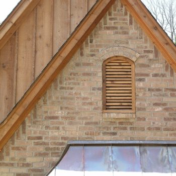 Gable end of a house with brick wall, wooden siding, and a small louvered vent window near the roof peak.