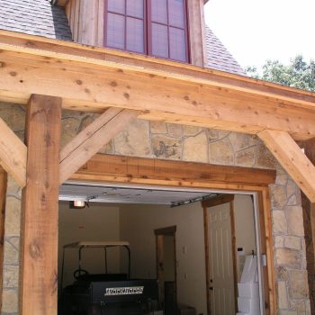 Wood beam garage entrance with stone facade, one door open showing interior and a vehicle inside, daylight scene.