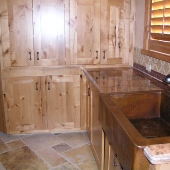 Wooden kitchen cabinets and drawers with a brown farmhouse sink, stone countertop, and tiled floor and backsplash.