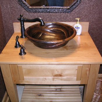 A bronze vessel sink with a black faucet on a wooden vanity, with soap dispenser and decorative mirror above.