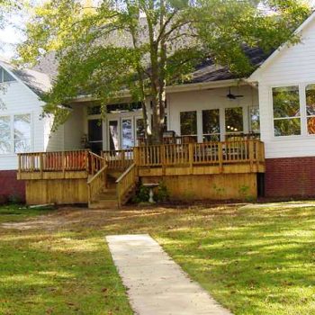 A white house with a brick foundation, wooden porch, and steps, set back from a sidewalk, surrounded by grass and trees.