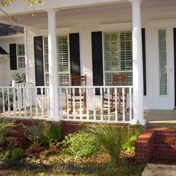 Front porch with white railing, black shutters, two wooden rocking chairs, and brick steps leading to a black front door.