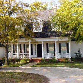 Two-story white house with black shutters, a front porch, and a curved driveway, surrounded by trees.