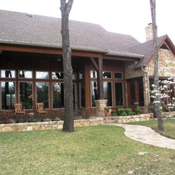 Backyard view of a stone and wood house with large windows, a covered porch, and a stone walkway on a grassy lawn.