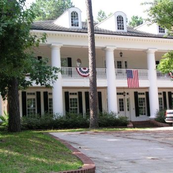 Large two-story house with white columns, American flag, and patriotic bunting, with a pickup truck in the driveway.