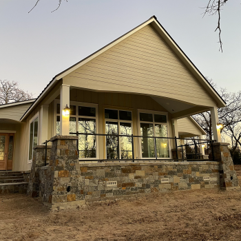 A modern house with stone and siding exterior, large windows, front porch, and steps, surrounded by bare trees and dirt ground.