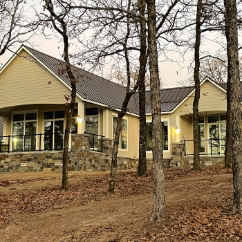 Single-story house with large windows and stone accents, surrounded by bare trees on a dirt and leaf-covered ground.