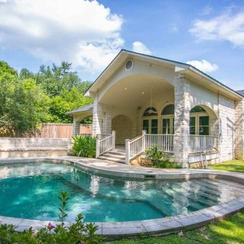 Stone house with arched patio next to a curved backyard swimming pool and surrounding greenery under a blue sky.