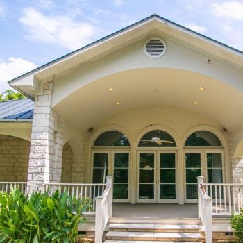 White house with stone columns, arched windows, a covered porch, railing, and surrounding greenery.
