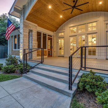 Modern house with a covered patio, large windows, railing, steps, and an American flag displayed on the side.