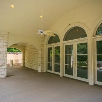 Covered patio with arched windows, a ceiling fan, and stone pillars, overlooking a fenced backyard.