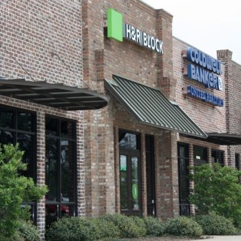 Brick storefronts with awnings, featuring H&R Block and Coldwell Banker signs, with bushes in front.