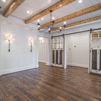 Empty room with wood ceiling beams, rustic sliding barn doors, wall sconces, and dark hardwood flooring. Kitchen in background.
