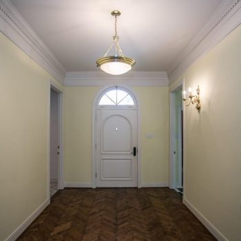 A hallway with light yellow walls, wood herringbone floor, white door with arched window, and ceiling lights.