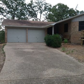 Single-story brick house with two-car garage, concrete driveway, and some shrubbery near the entrance.