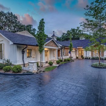 Single-story white house with a dark roof, large driveway, and landscaped yard at dusk with trees and sunset sky.