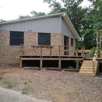 Single-story brick house with a new wooden deck and stairs, surrounded by trees and unpaved ground.