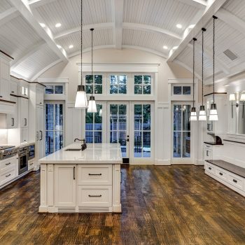 Spacious modern kitchen with white cabinets, pendant lights, large island, and vaulted beamed ceiling on wooden flooring.