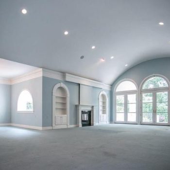 Spacious empty living room with vaulted ceiling, large arched windows, built-in shelves, and a fireplace.