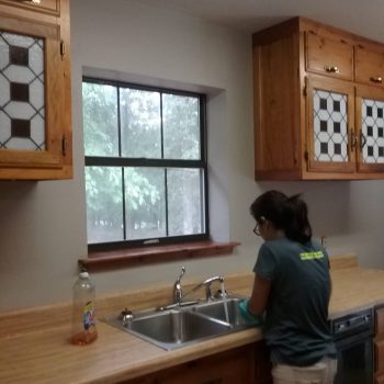 Person washing dishes at a kitchen sink with wooden cabinets and a large window overlooking trees.