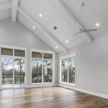 Bright empty room with vaulted ceiling, large windows, wood floors, and ceiling fan, overlooking trees.