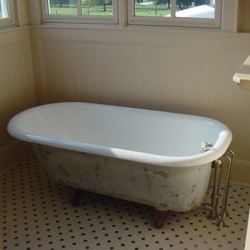 A clawfoot bathtub with chipped paint stands on a tiled floor in a sunlit bathroom corner.