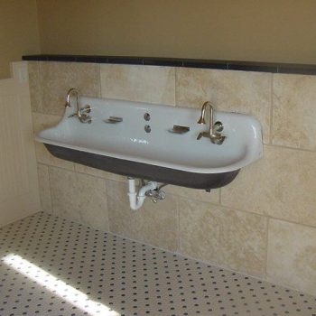 A double faucet wall-mounted sink with exposed plumbing in a tiled bathroom with a patterned floor.