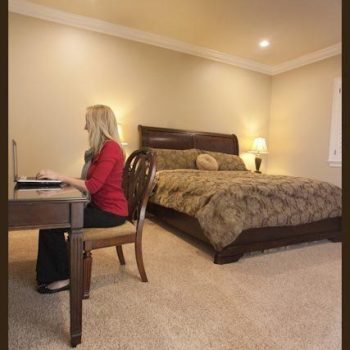 A woman sits at a desk using a laptop in a carpeted bedroom with a large bed and bedside lamp.